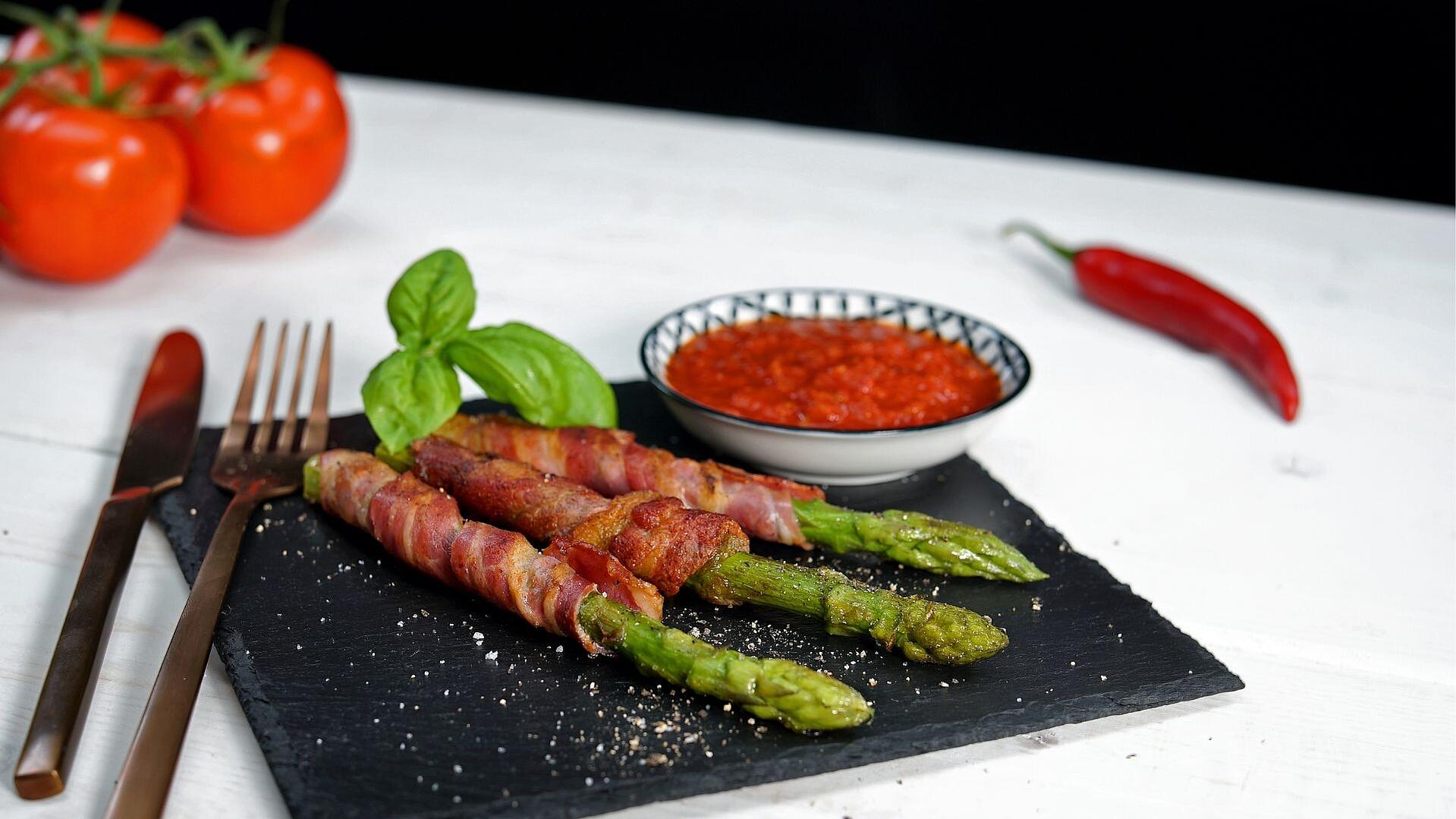 Grüner Spargel im Speckmantel angerichtet auf einer Schieferplatte mit Tomaten Sugo