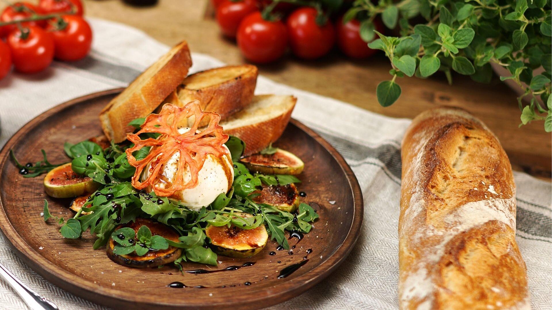 Auf einem Holzteller ist ein Salat mit Burrata und Feigen sowie Ciabatta Brot angerichtet.
