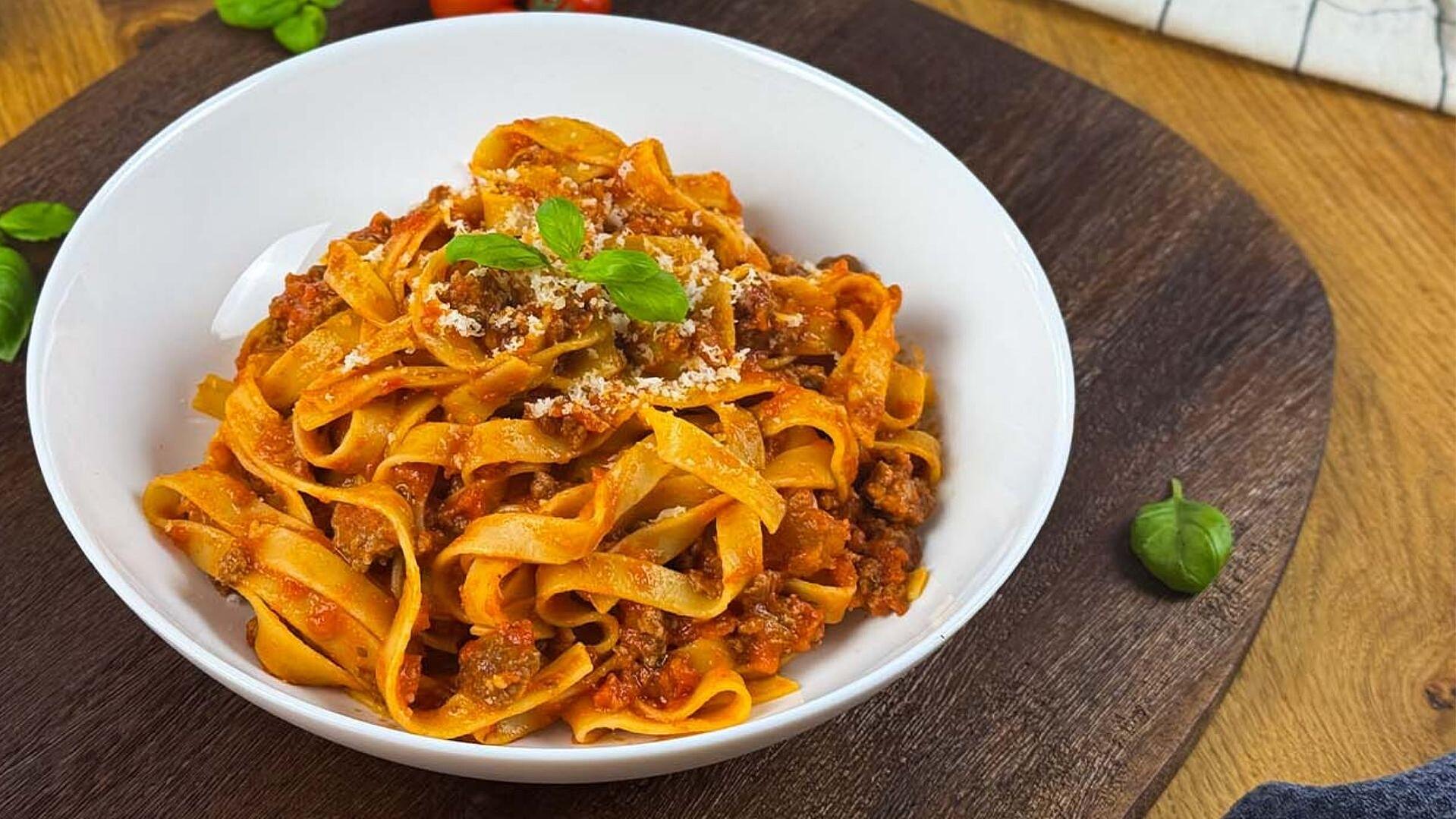 Ein Teller mit frischer Pasta und Bolognese, garniert mit Basilikumblättern, serviert auf einem rustikalen Holzbrett.